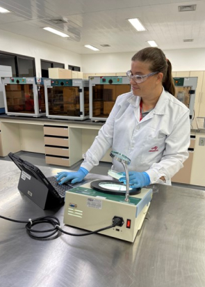 Imagem de uma cientista fazendo um teste com um frasco e conferindo os resultados em um notebook, ao fundo há dispensas de amostras e gavetas em um laboratório